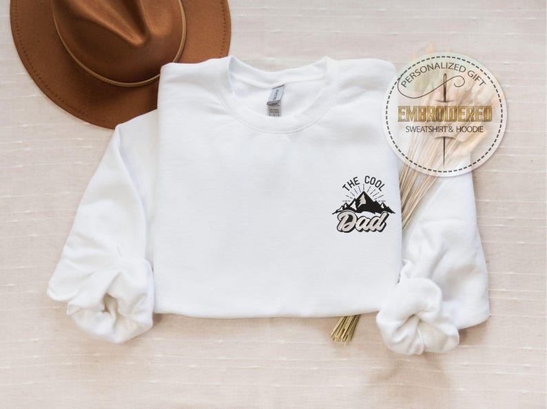Op de afbeelding: Witte sweatshirt met de tekst "The Cool Dad" in het zwart, met een bergafbeelding. Op de achtergrond is een bruine hoed te zien. De sweatshirt heeft lange mouwen en een ronde hals. Een ronde sticker luidt "Gepersonaliseerd cadeau, Geborduurd sweatshirt & hoodie."
