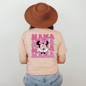 以下が含まれることがあります： 桃色のTシャツに、ピンクとパープルのミニーマウスのグラフィックがプリントされています。"MAMA"という文字が4回繰り返されています。