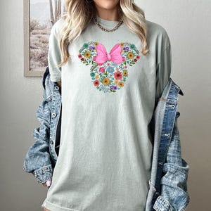 Op de afbeelding: Lichtgroen t-shirt met een bloemen Minnie Mouse-ontwerp. Het ontwerp bevat een roze strik en kleurrijke bloemen in een Mickey Mouse-silhouet. Het shirt is een comfortabele, casual stijl.