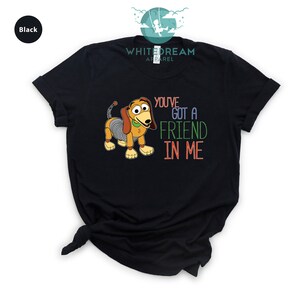 Op de afbeelding: Zwart T-shirt met een cartoon afbeelding van een hondenspeelgoed uit de Toy Story-filmfranchise. De tekst "You've got a friend in me" staat in kleurrijke letters op het shirt gedrukt.