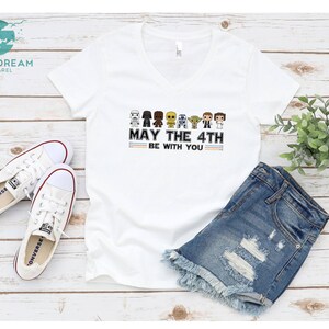 Op de afbeelding: Wit V-hals T-shirt met een afbeelding van Star Wars-personages en de tekst "May the 4th Be With You". Het shirt wordt getoond op een houten oppervlak met een paar witte sneakers en een spijkerbroek.