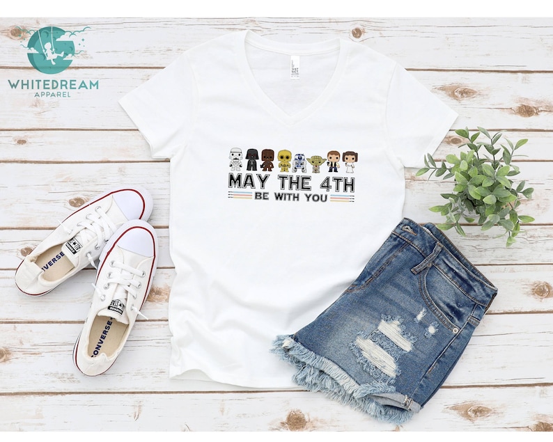 Op de afbeelding: Wit V-hals T-shirt met een afbeelding van Star Wars-personages en de tekst "May the 4th Be With You". Het shirt wordt getoond op een houten oppervlak met een paar witte sneakers en een spijkerbroek.