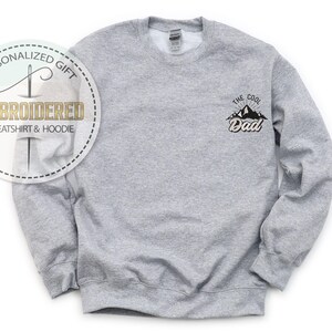 Op de afbeelding: Een grijze sweatshirt met de tekst "THE COOL Dad" en een bergafbeelding op de linkerbovenborst. Een cirkelvormige afbeelding linksboven leest "PERSONALIZED GIFT EMBROIDERED SWEATSHIRT & HOODIE".