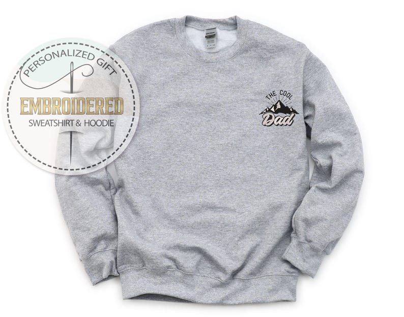 Op de afbeelding: Een grijze sweatshirt met de tekst "THE COOL Dad" en een bergafbeelding op de linkerbovenborst. Een cirkelvormige afbeelding linksboven leest "PERSONALIZED GIFT EMBROIDERED SWEATSHIRT & HOODIE".
