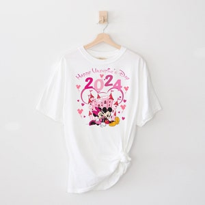 May include: White t-shirt with a pink and white graphic of Mickey Mouse and Minnie Mouse in front of a pink castle. The graphic also includes the text "Happy Valentine's Day 2024" with pink hearts.
