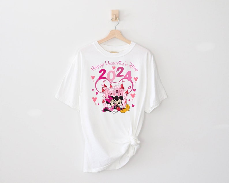 May include: White t-shirt with a pink and white graphic of Mickey Mouse and Minnie Mouse in front of a pink castle. The graphic also includes the text "Happy Valentine's Day 2024" with pink hearts.
