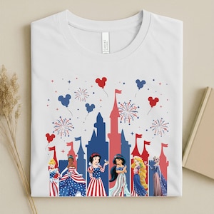 K&ouml;nnte beinhalten: Wei&szlig;es T-Shirt mit einem patriotischen Design. Es zeigt eine rote und blaue Schloss-Silhouette, Feuerwerk und Disney-Prinzessinnen-Charaktere. Die Prinzessinnen sind in rot, wei&szlig; und blau gekleidet, mit Mickey-Mouse-f&ouml;rmigen Ballons.