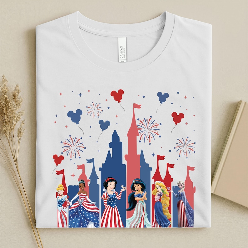 K&ouml;nnte beinhalten: Wei&szlig;es T-Shirt mit einem patriotischen Design. Es zeigt eine rote und blaue Schloss-Silhouette, Feuerwerk und Disney-Prinzessinnen-Charaktere. Die Prinzessinnen sind in rot, wei&szlig; und blau gekleidet, mit Mickey-Mouse-f&ouml;rmigen Ballons.