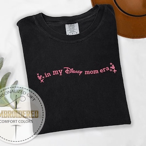 May include: Black t-shirt with pink embroidered text that says "in my Disney mom era" with two pink Mickey Mouse heads on either side of the text.