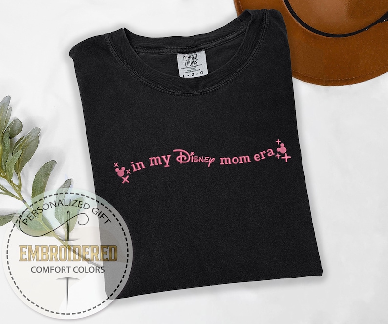 May include: Black t-shirt with pink embroidered text that says "in my Disney mom era" with two pink Mickey Mouse heads on either side of the text.