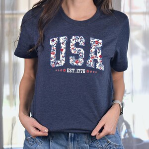 May include: Navy blue t-shirt with the word "USA" in a floral design. The shirt also features the text "EST. 1776" with stars. The t-shirt is a crew neck style.