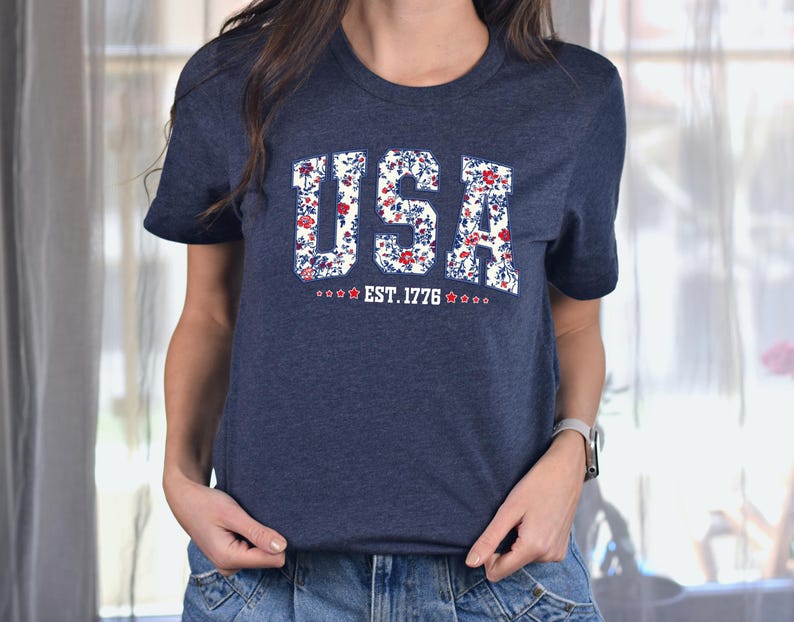 May include: Navy blue t-shirt with the word "USA" in a floral design. The shirt also features the text "EST. 1776" with stars. The t-shirt is a crew neck style.