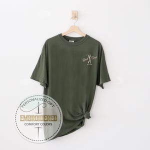 K&ouml;nnte beinhalten: Olivgr&uuml;nes T-Shirt mit dem Text "Best Dad" und einer Golfschl&auml;ger-Grafik. Das Shirt h&auml;ngt an einem Holzb&uuml;gel vor wei&szlig;em Hintergrund. Ein kreisf&ouml;rmiges Logo mit den Worten "Personalized Gift" und "Embroidered Comfort Colors" ist sichtbar.