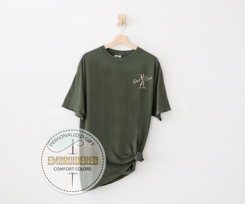 K&ouml;nnte beinhalten: Olivgr&uuml;nes T-Shirt mit dem Text "Best Dad" und einer Golfschl&auml;ger-Grafik. Das Shirt h&auml;ngt an einem Holzb&uuml;gel vor wei&szlig;em Hintergrund. Ein kreisf&ouml;rmiges Logo mit den Worten "Personalized Gift" und "Embroidered Comfort Colors" ist sichtbar.