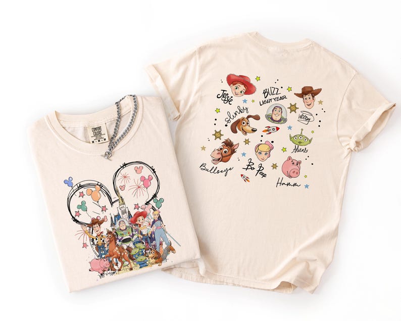 May include: Two cream-colored t-shirts. One features a Mickey Mouse head design with Disney characters. The other shirt has various Toy Story characters and text. A silver chain necklace is draped over the first shirt.