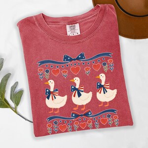 May include: A red Comfort Colors t-shirt featuring a patriotic design with three white ducks wearing bow ties with red, white, and blue accents. The shirt also has decorative ribbons, hearts, and floral elements.