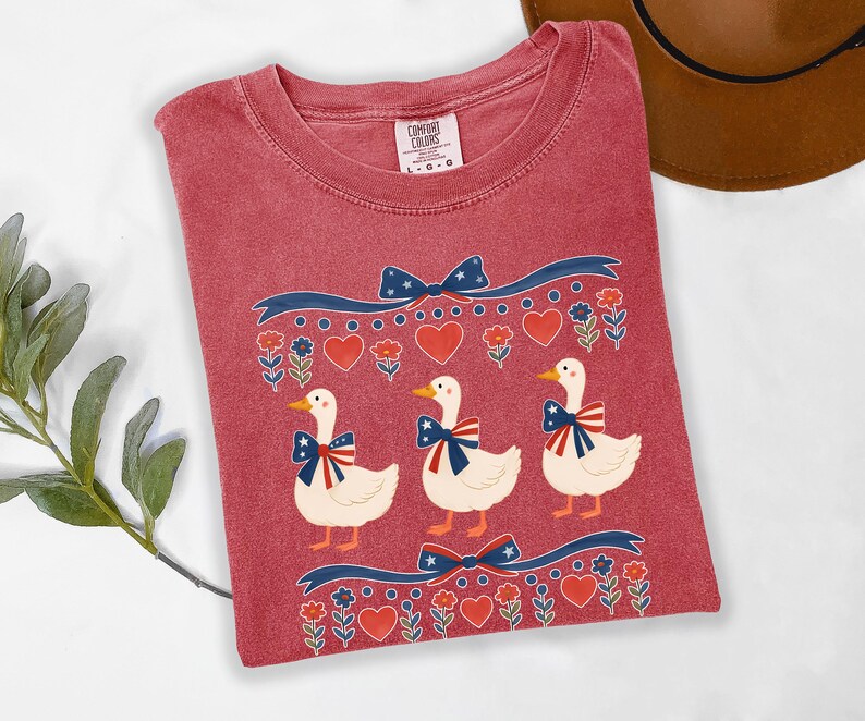 May include: A red Comfort Colors t-shirt featuring a patriotic design with three white ducks wearing bow ties with red, white, and blue accents. The shirt also has decorative ribbons, hearts, and floral elements.