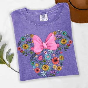 Op de afbeelding: Een paars Comfort Colors T-shirt met een bloemenontwerp in de vorm van een strik en muisoren. Het ontwerp bevat een grote roze strik en kleurrijke bloemen. Het shirt ligt opgevouwen op een wit oppervlak.