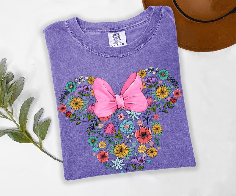Op de afbeelding: Een paars Comfort Colors T-shirt met een bloemenontwerp in de vorm van een strik en muisoren. Het ontwerp bevat een grote roze strik en kleurrijke bloemen. Het shirt ligt opgevouwen op een wit oppervlak.