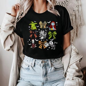May include: Black t-shirt featuring colorful bow designs with characters from a popular animated movie. The bows include characters like Sally, Jack Skellington, and Oogie Boogie. The shirt has a crew neck and short sleeves.