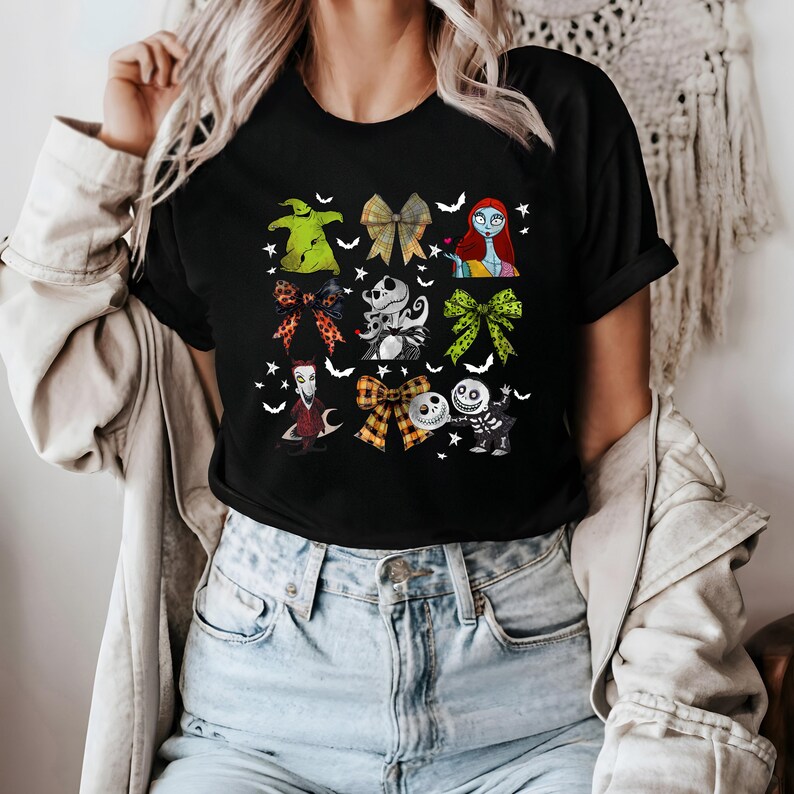 May include: Black t-shirt featuring colorful bow designs with characters from a popular animated movie. The bows include characters like Sally, Jack Skellington, and Oogie Boogie. The shirt has a crew neck and short sleeves.