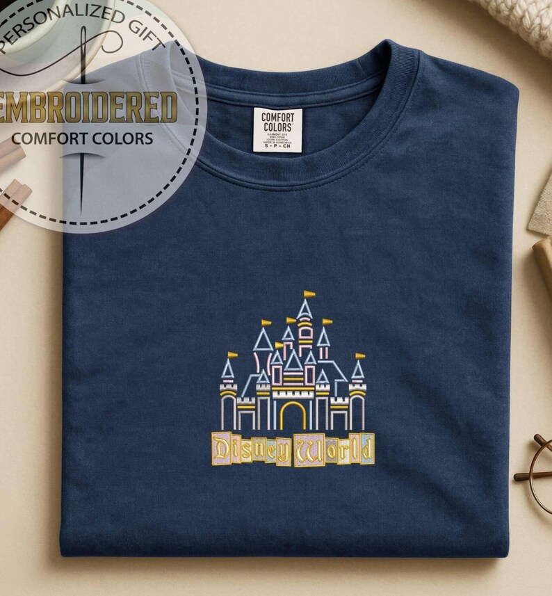 May include: Navy blue t-shirt with an embroidered Disney World castle design in yellow, white, and blue. The shirt has a crew neck and short sleeves. The text "Disney World" is embroidered below the castle.
