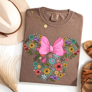 Op de afbeelding: Een bruin T-shirt met een bloemen Minnie Mouse-ontwerp en een grote roze strik. Het ontwerp is omgeven door kleurrijke bloemen. Een gouden ketting, een beige hoed en bruine sandalen staan ook op de foto.