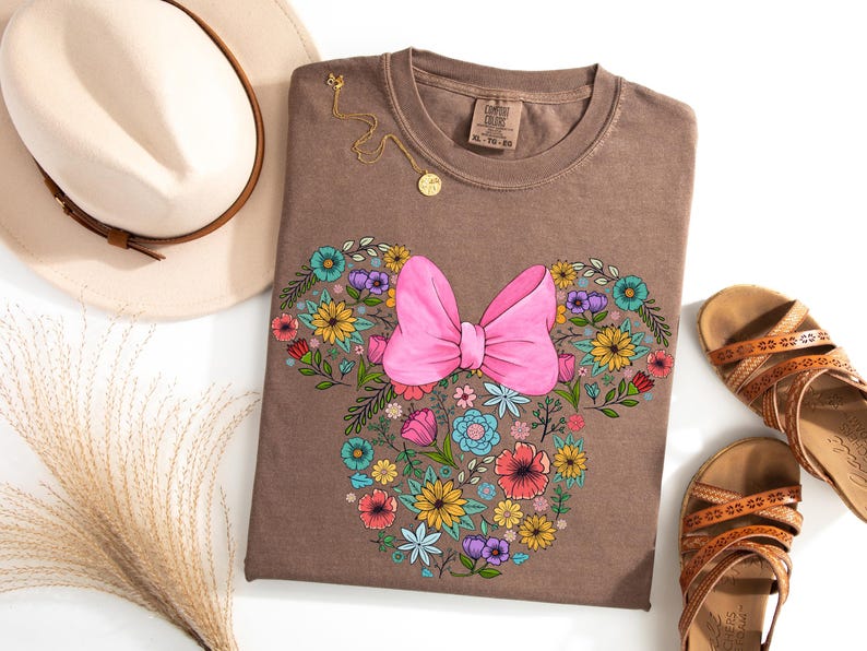 Op de afbeelding: Een bruin T-shirt met een bloemen Minnie Mouse-ontwerp en een grote roze strik. Het ontwerp is omgeven door kleurrijke bloemen. Een gouden ketting, een beige hoed en bruine sandalen staan ook op de foto.