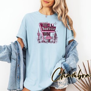 May include: Light blue t-shirt with a graphic of a retro motel sign that reads "Welcome to Read On Bookville".
