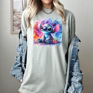 Pu&ograve; includere: Una maglietta verde chiaro con un'illustrazione ad acquerello di Stitch di Lilo & Stitch. L'opera d'arte mostra Stitch su uno sfondo colorato di tonalit&agrave; rosa, blu e viola. La maglietta &egrave; uno stile casual e comodo.