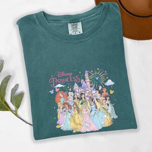 May include: Teal-colored t-shirt featuring a graphic of Disney Princesses in colorful gowns, with a castle and fireworks. The words "Disney Princess" are written in pink script. The shirt is made of a soft material.