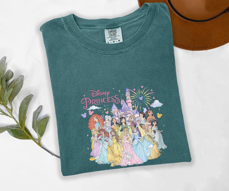 May include: Teal-colored t-shirt featuring a graphic of Disney Princesses in colorful gowns, with a castle and fireworks. The words "Disney Princess" are written in pink script. The shirt is made of a soft material.