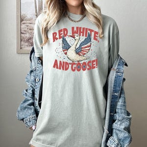 May include: A light green t-shirt with the text "RED, WHITE, AND GOOSE!" in red letters. The graphic features a goose with wings designed like the American flag. The shirt is worn with a denim jacket.