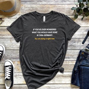 May include: A dark gray t-shirt with white text that reads "IF YOU'VE EVER WONDERED WHAT YOU WOULD HAVE DONE IN 1930s GERMANY: You are doing it right now."