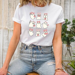 Puede incluir: Camiseta blanca con un gr&aacute;fico de nueve fantasmas lindos con lazos rosas y sosteniendo varios art&iacute;culos. Los fantasmas est&aacute;n dispuestos en un patr&oacute;n de cuadr&iacute;cula.