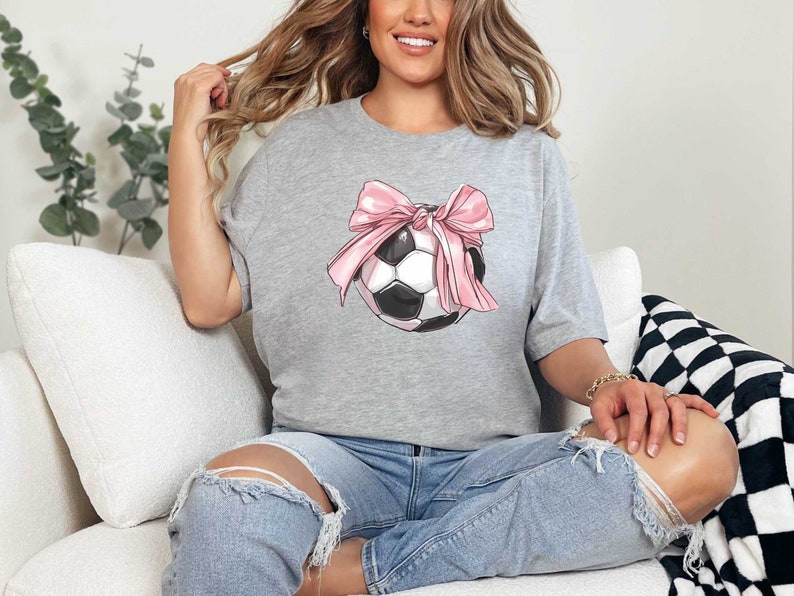 May include: A gray t-shirt with a graphic of a soccer ball with a pink bow on it.