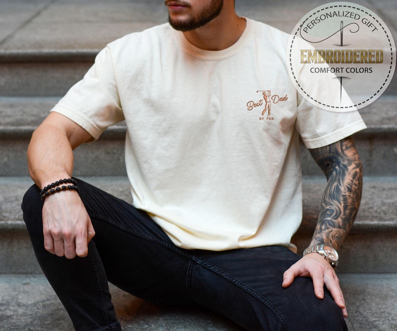 K&ouml;nnte beinhalten: Cremefarbenes T-Shirt mit dem gestickten Text "Best Dad by Par" und einer Golf-Grafik. Das Shirt hat auch ein rundes Emblem mit der Aufschrift "Personalized Gift" und "Embroidered Comfort Colors."