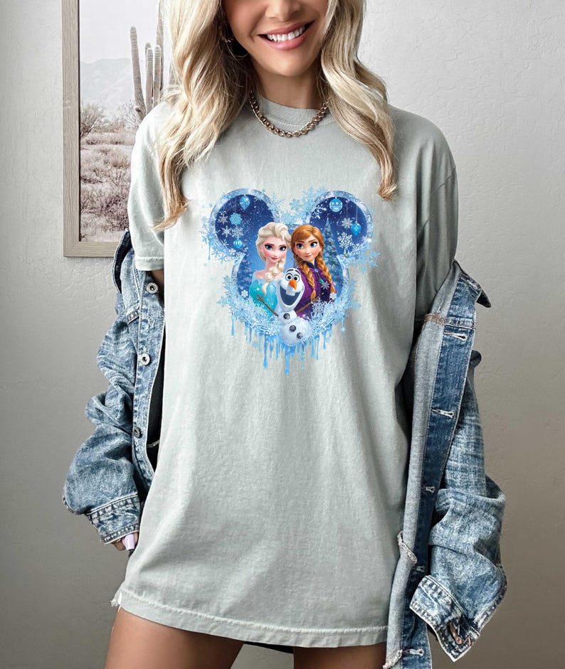 Puede incluir: Camiseta verde claro con un gr&aacute;fico de los personajes de Frozen de Disney, Elsa, Anna y Olaf, dentro de un contorno de la cabeza de Mickey Mouse. El dise&ntilde;o incluye copos de nieve y detalles en azul hielo. La camiseta es de estilo casual.