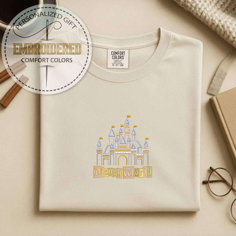 May include: Cream-colored sweatshirt with an embroidered Disney World castle design in yellow, blue, and pink. The text "Disney World" is embroidered below the castle. The sweatshirt has a Comfort Colors tag.