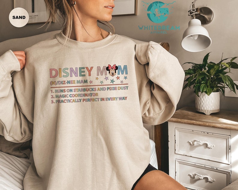 May include: A beige sweatshirt with a colorful graphic that says "Disney Mom" with a Minnie Mouse illustration. The text below the graphic says "(N)/DIZ-NEE MAM" and lists three reasons why a Disney Mom is the best.