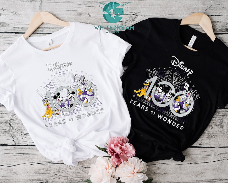 May include: Two t-shirts, one white and one black, with a graphic of Mickey Mouse, Minnie Mouse, Donald Duck, Goofy, and Pluto celebrating Disney's 100th anniversary. The graphic features the text "Disney 100 Years of Wonder".