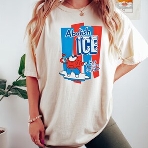 May include: Cream-colored t-shirt with a graphic that reads "Abolish ICE" in blue and red. The graphic features a cartoon polar bear holding a bottle. The text "keep families together" is also included.