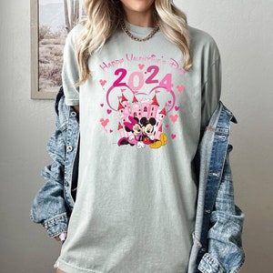 May include: A light green t-shirt with a pink graphic of Mickey Mouse and Minnie Mouse in front of a pink castle. The graphic also includes the text "Happy Valentine's Day 2024" in pink.