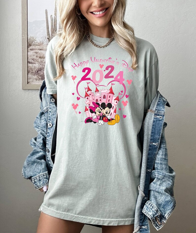 May include: A light green t-shirt with a pink graphic of Mickey Mouse and Minnie Mouse in front of a pink castle. The graphic also includes the text "Happy Valentine's Day 2024" in pink.
