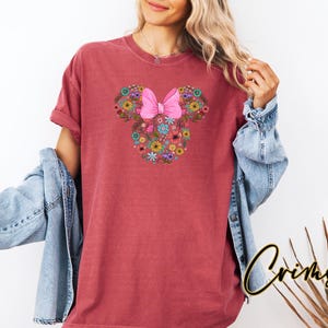 Op de afbeelding: Een bordeauxrood T-shirt met een bloemen Minnie Mouse-ontwerp en een roze strik. Het ontwerp is gemaakt van diverse kleurrijke bloemen. Het shirt wordt gedragen met een spijkerjasje.