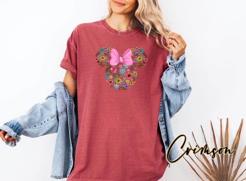 Op de afbeelding: Een bordeauxrood T-shirt met een bloemen Minnie Mouse-ontwerp en een roze strik. Het ontwerp is gemaakt van diverse kleurrijke bloemen. Het shirt wordt gedragen met een spijkerjasje.
