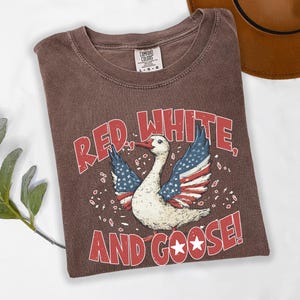 May include: Brown t-shirt with a distressed graphic of a goose with American flag-themed wings and the text "RED, WHITE, AND GOOSE!". The shirt has a crew neck and a Comfort Colors tag.