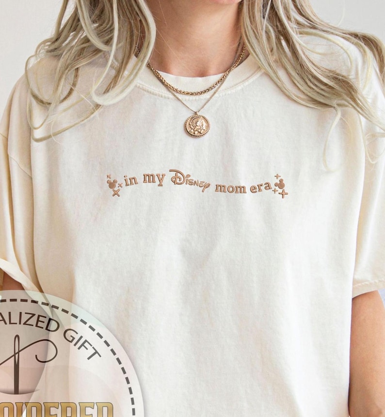May include: A white t-shirt with the text "in my Disney mom era" in brown lettering. The shirt features a small Mickey Mouse graphic and stars.