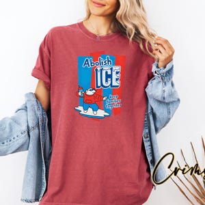 May include: A red t-shirt with a graphic that says "Abolish ICE" in blue and white, with a cartoon polar bear holding an ice pop. The text "keep families together" is also included. The shirt is worn by a person.
