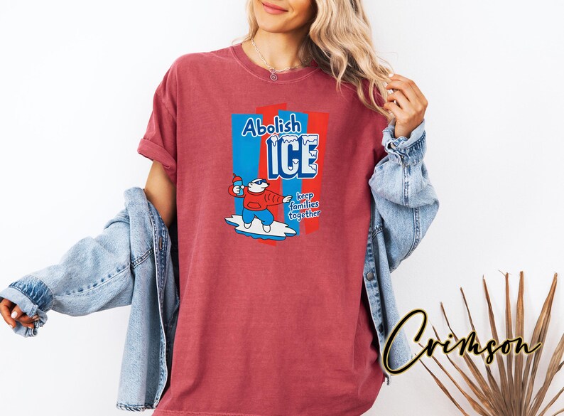 May include: A red t-shirt with a graphic that says "Abolish ICE" in blue and white, with a cartoon polar bear holding an ice pop. The text "keep families together" is also included. The shirt is worn by a person.